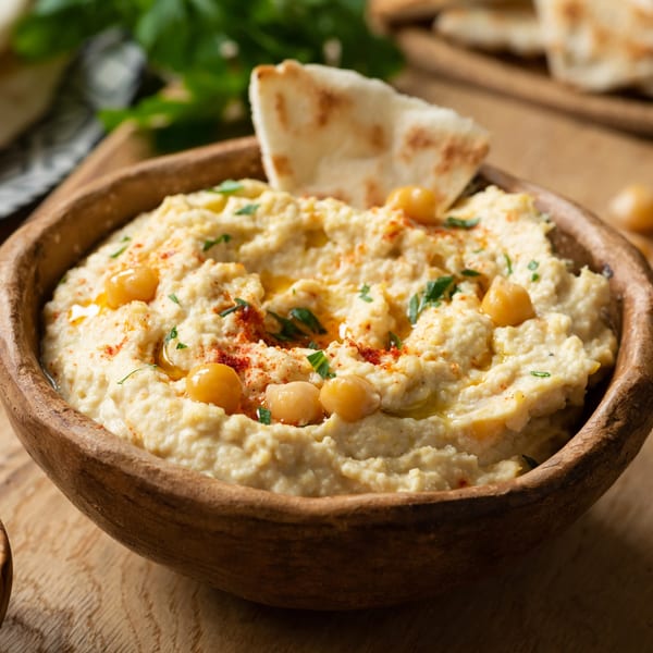 A wooden bowl of hummus, garnished with chickpeas, paprika, olive oil, and parsley, with pita bread.