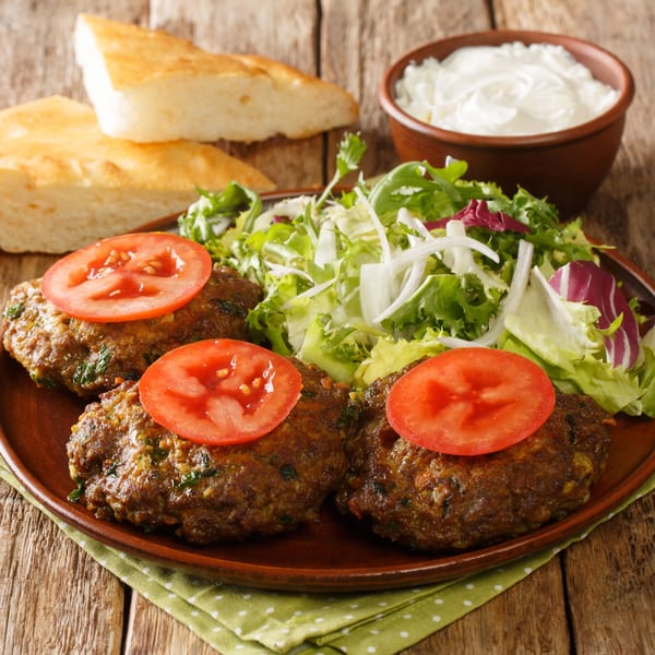 Three Mediterranean kebab patties topped with tomato slices, served with fresh salad, bread, and yogurt.