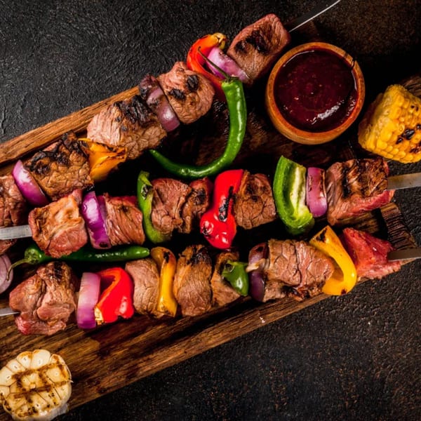 Grilled beef and vegetable kebabs with sauce, corn, and garlic on a wooden board.