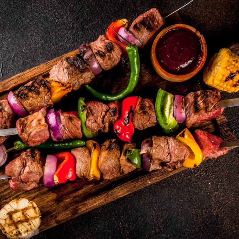 Grilled beef and vegetable kebabs with sauce, corn, and garlic on a wooden board.