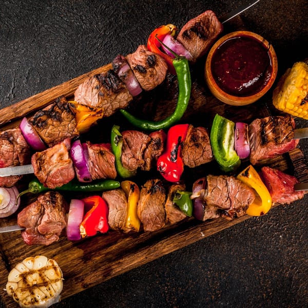 Grilled beef and vegetable kebabs on skewers, with sauce, corn, and garlic on a wooden board.