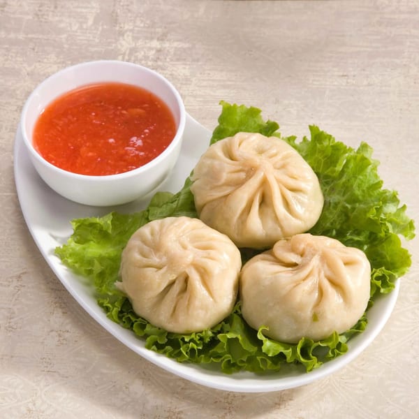 Three steamed momos on lettuce with a side of red dipping sauce on a white plate.