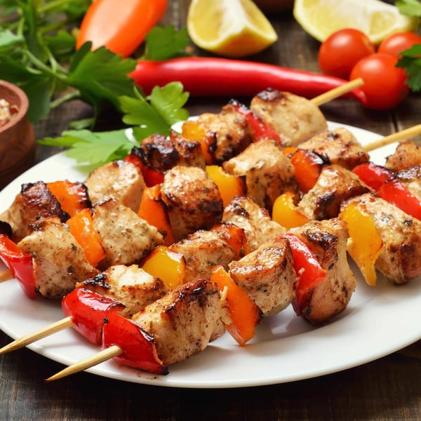 Grilled chicken and bell pepper kebabs on a white plate, with fresh vegetables in the background.