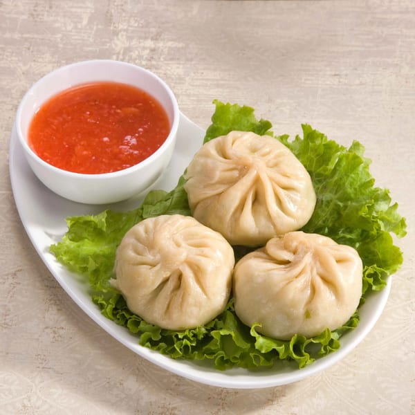 Three steamed momos on lettuce with a side of red dipping sauce on a white plate.