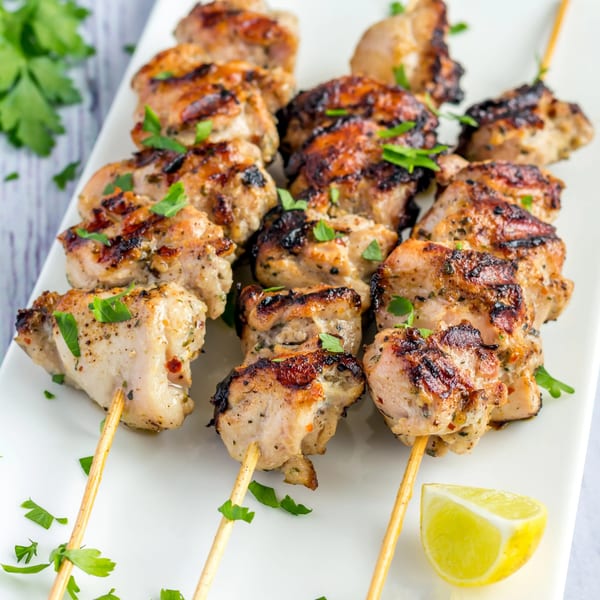 Grilled chicken skewers with parsley and a lemon wedge on a white plate.