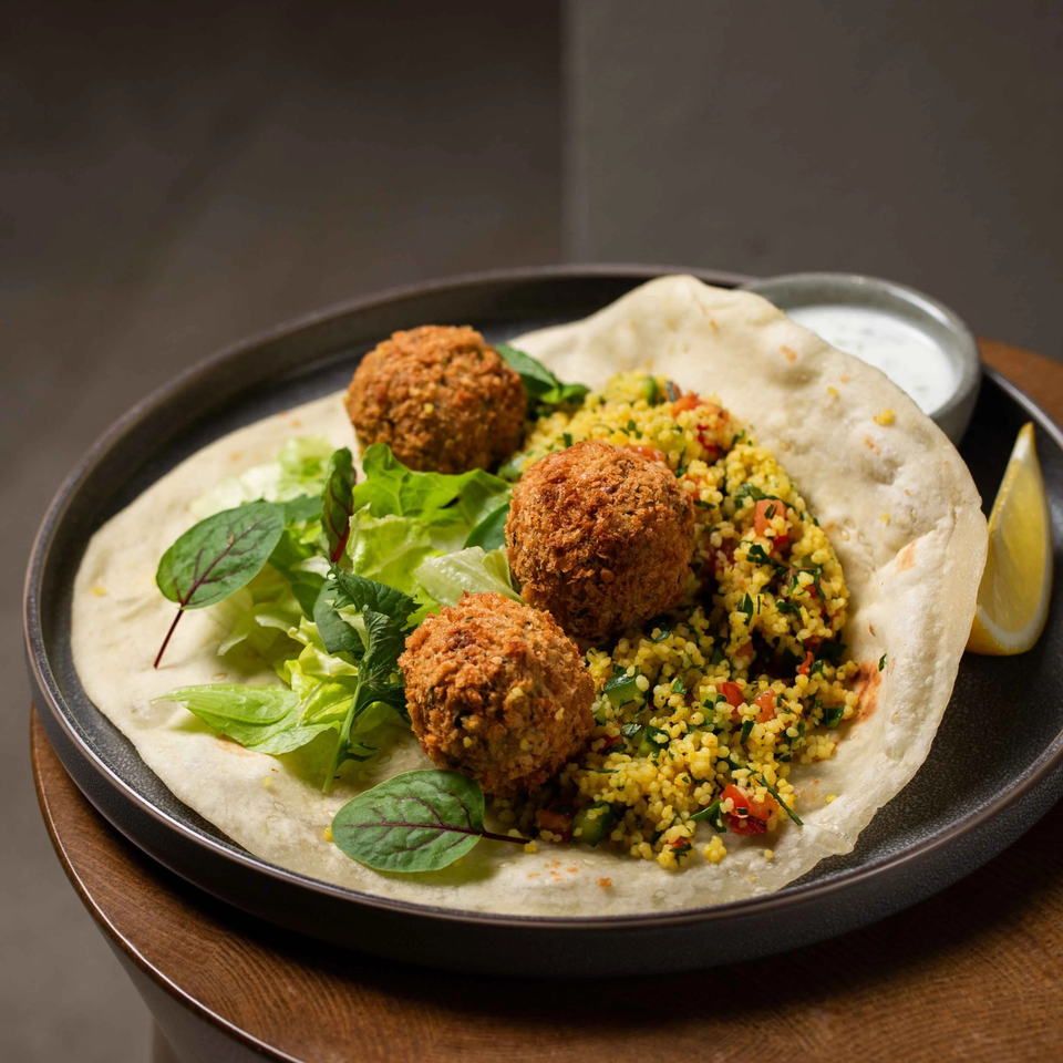 Mediterranean falafel wrap with couscous, fresh greens, a lemon wedge, and white sauce.