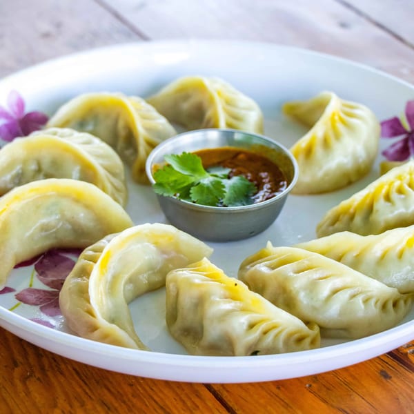A white plate with Kebab & MO:MO's steamed momos and a small metal bowl of dipping sauce.